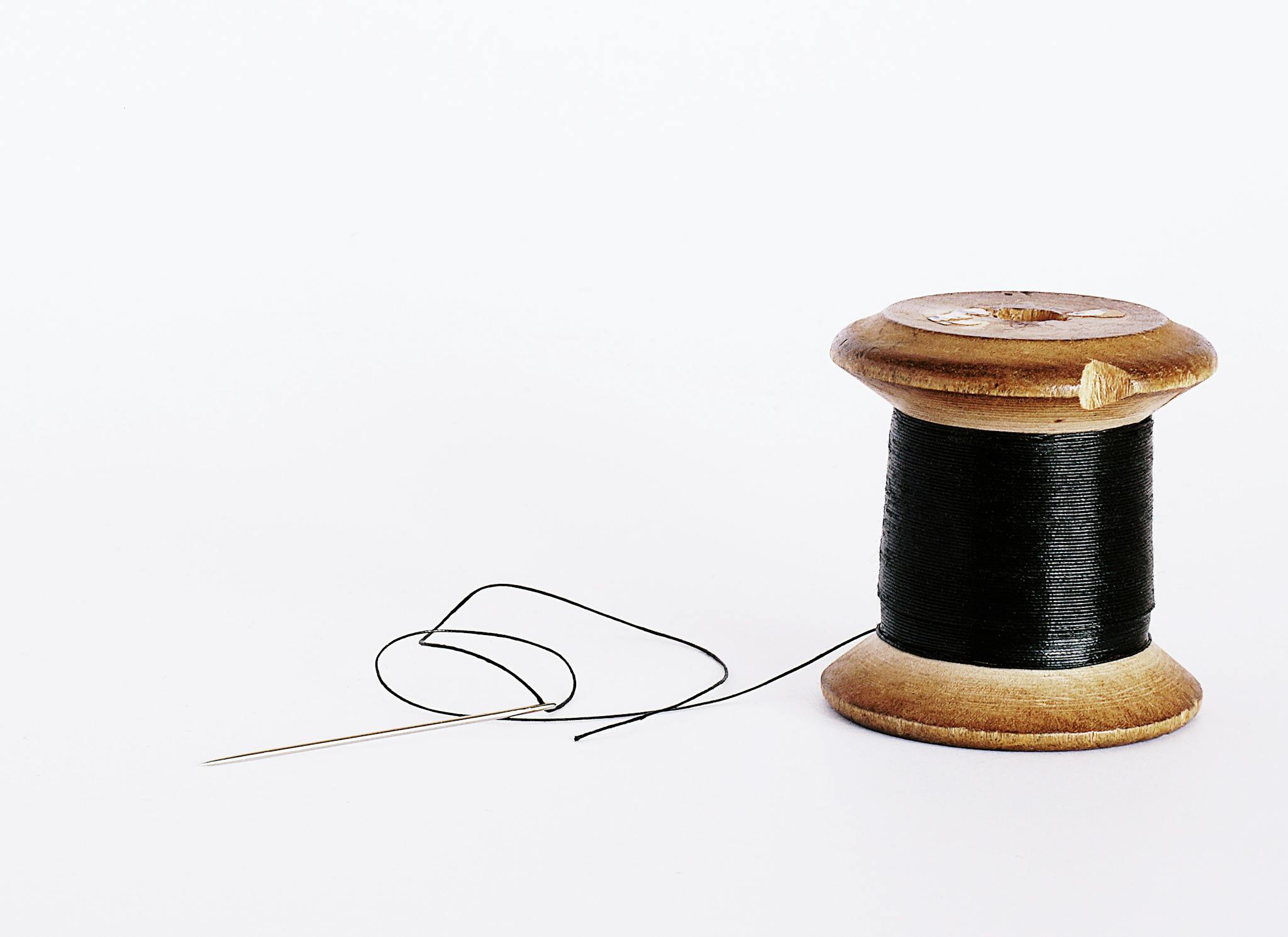 Close-up of a wooden spool with black thread and a sewing needle on a white background.